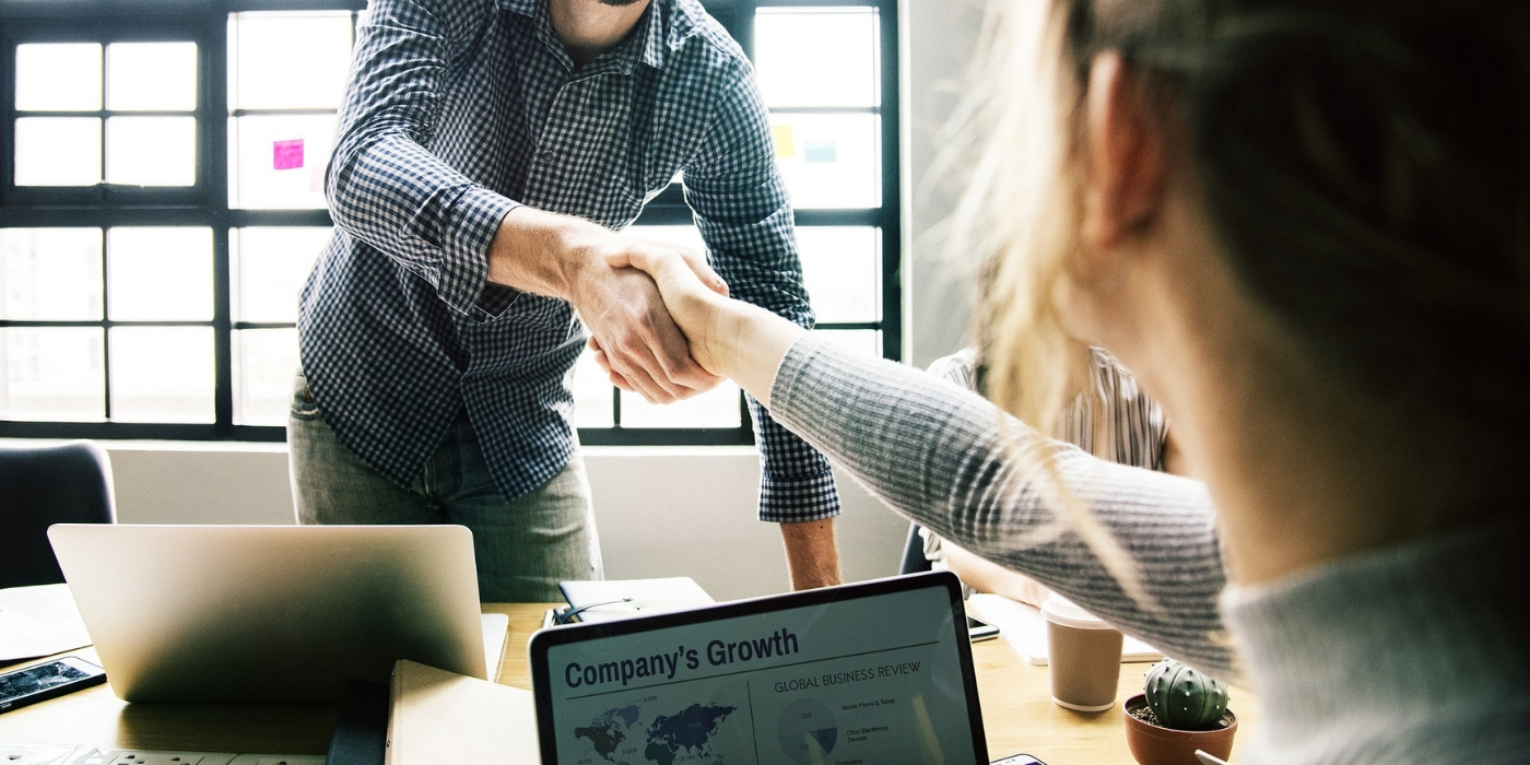 man in checkered shirt shaking the hand of a woman in a grey sweater who is at a laptop