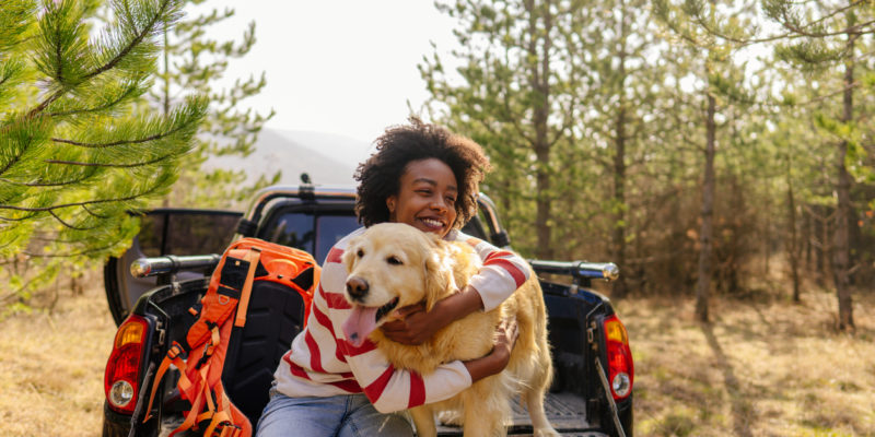 woman smiling and hugging a panting golden retriever next to an orange backpack in the bed of a pickup truck
