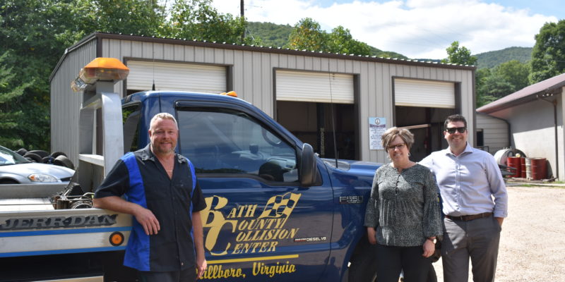 Two men and a woman stand next to a blue truck with a yellow Bath County Collision Center logo on the side