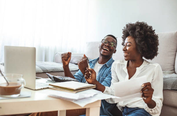 woman and man sit on the floor in front of their couch laughing, holding papers, and using a laptop