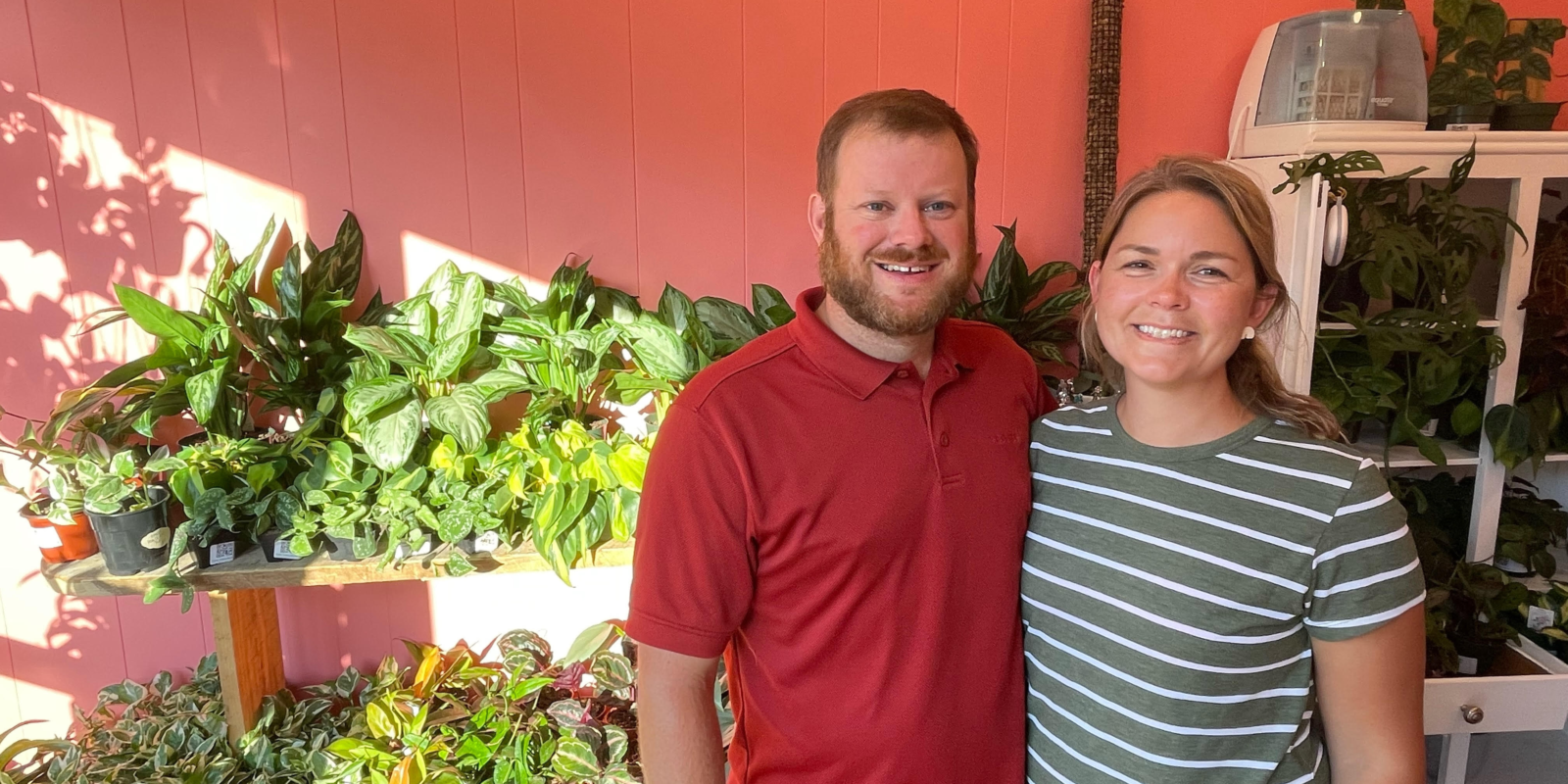 Man in red shirt and woman in green striped shirt smile and stand in front of a shelf of plants