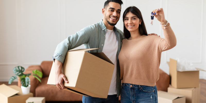 man holding a cardboard box and woman holding a pair of keys smile in front of their brown couch