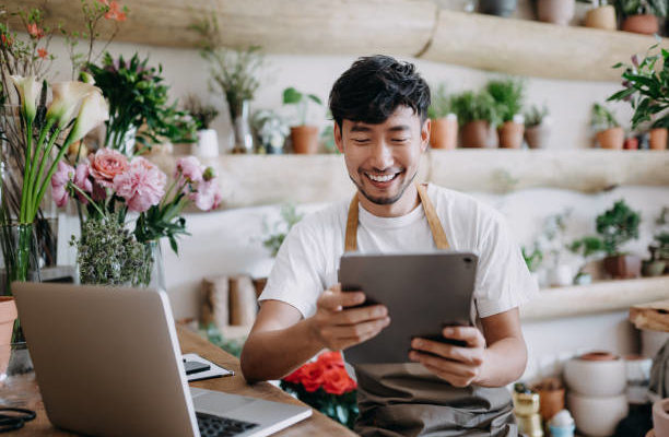 man in an apron sits in a plant shop in front of a laptop, smiling and holding an ipad