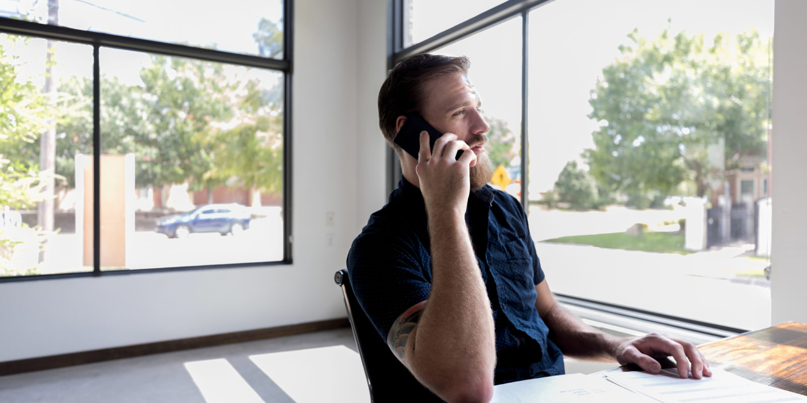 man in a blue button down short sleeve sits in front of windows and talks on a smartphone