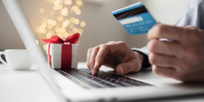 person using a laptop holds a blue credit card with a tiny christmas present and twinkling christmas lights in the background