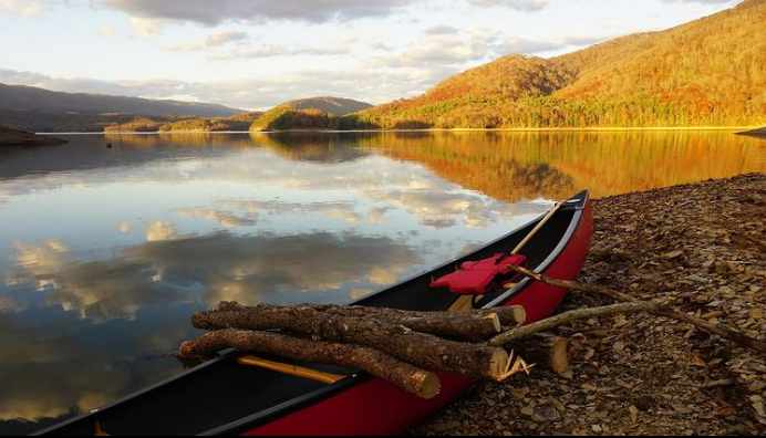 Canoe laden with pieces of lumber sits on the shore of a glassy lake that reflects the sky above and the mountains beyond