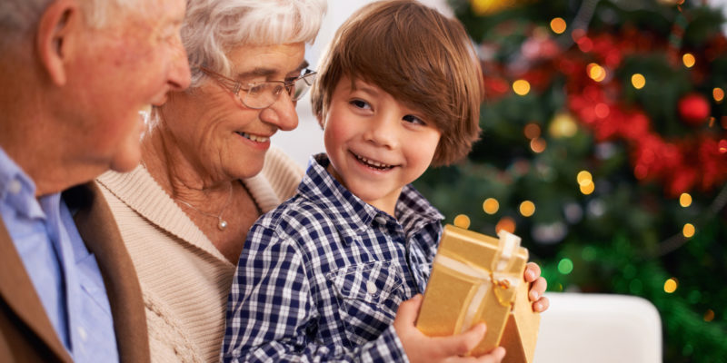 Brown-haired boy sitting with grandparents and holding a present wrapped in yellow paper in front of a christmas tree