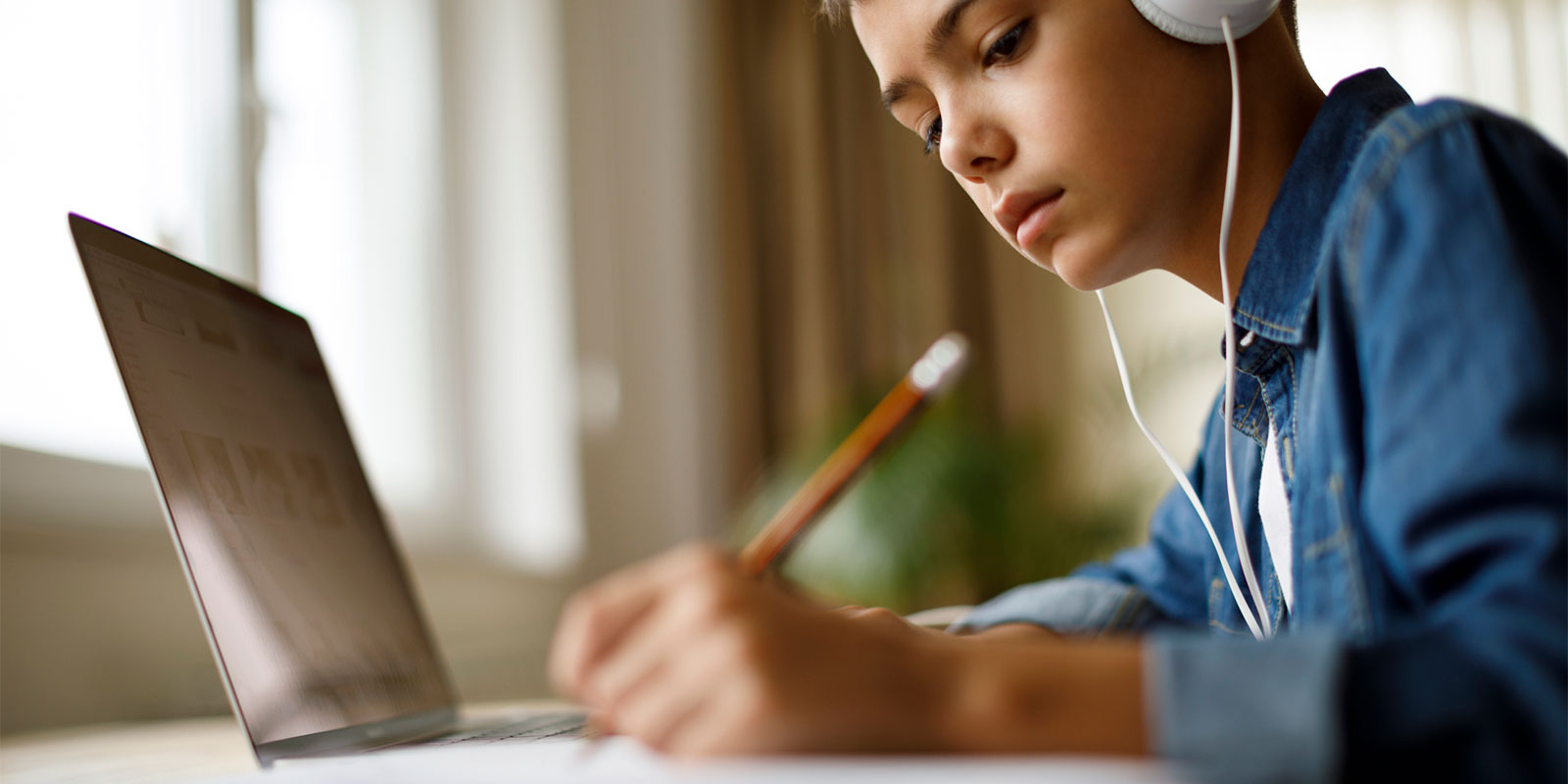 young boy in a blue shirt and headphones sits at a computer and writes with a pencil