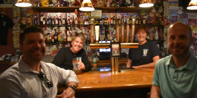 Two men in business shirts in front of a bar and two female employees behind the bar smile at the camera