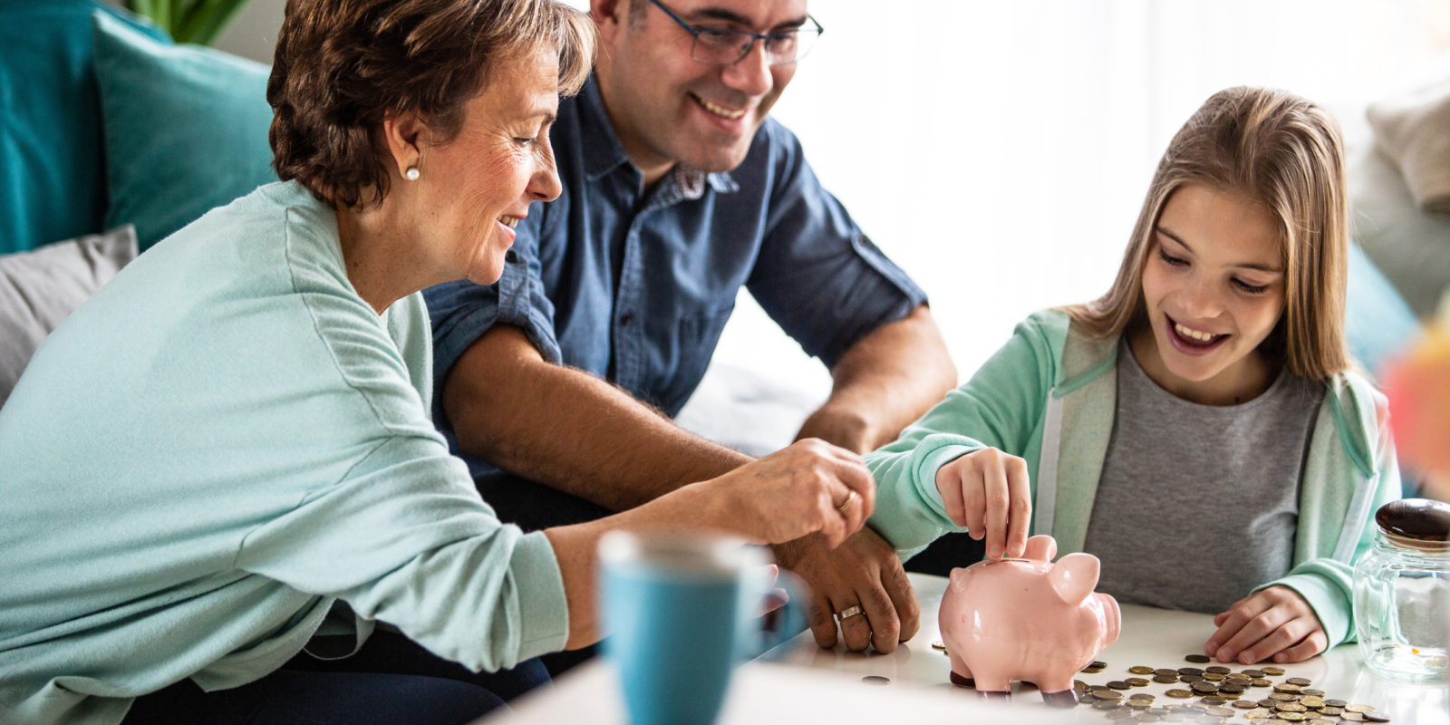 Blonde girl in green zip up sweatshirt sits at a table with a man and a woman and sticks coins into a pink piggy bank