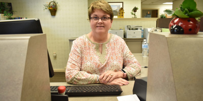 woman in a green and orange paisley shirt and glasses sits behind a desk and smiles