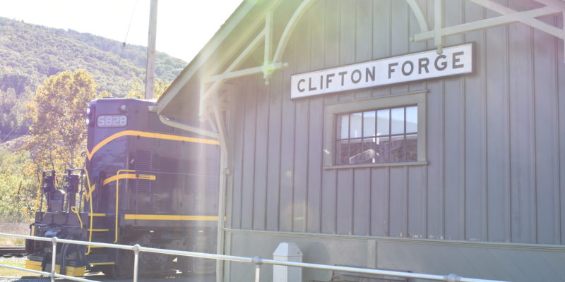 Gray green exterior of Clifton Forge train station with a black and yellow train car in the background