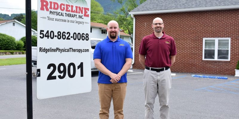 Two men stand in front of a brick building next to a sign that reads "Ridgeline Physical Therapy, Inc.: Your success is our legacy. 540-862-0068, ridgelinephysicaltherapy.com, 2901"