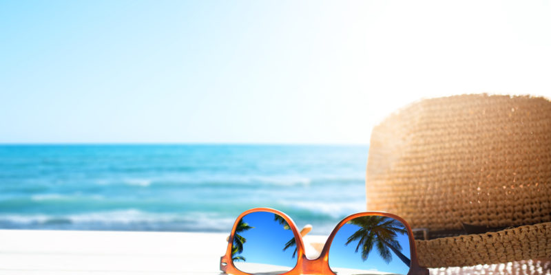 a beach hat and a pair of orange sunglasses reflecting palm trees with an ocean in the background