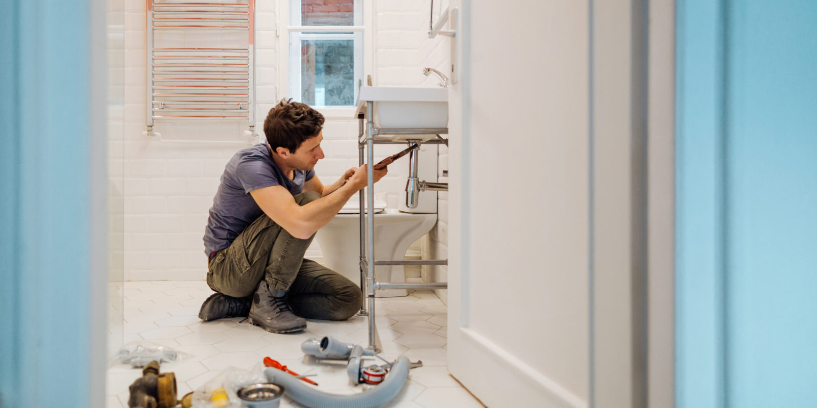 brown-haired man in a blue shirt and olive pants fixing the pipe under a bathroom sink