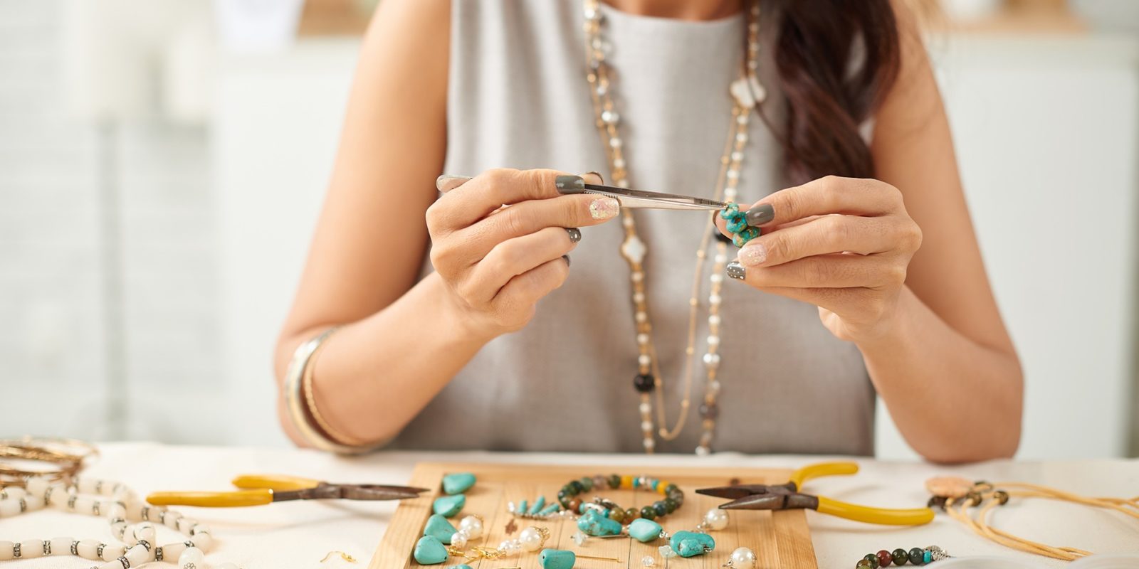woman in gray tank top and golden necklaces uses a jewelers tool to assemble jewelery at a workstation