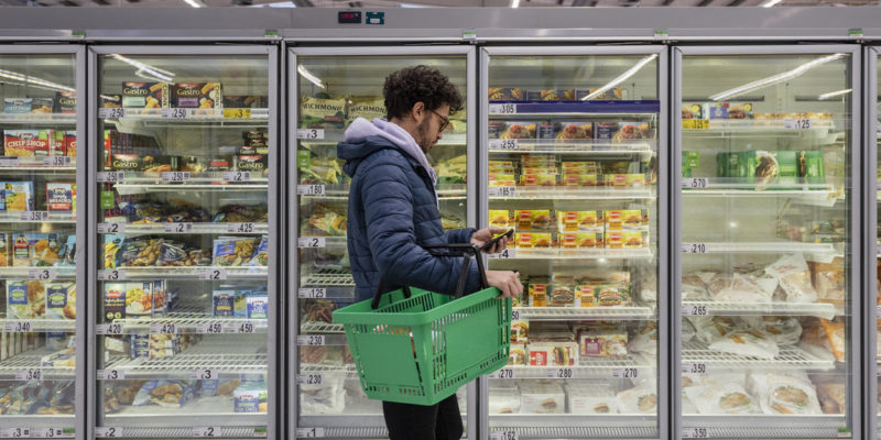 man in a puffy blue jacket and purple hoodie and holding a green shopping basket shopping in the frozen section of a grocery store