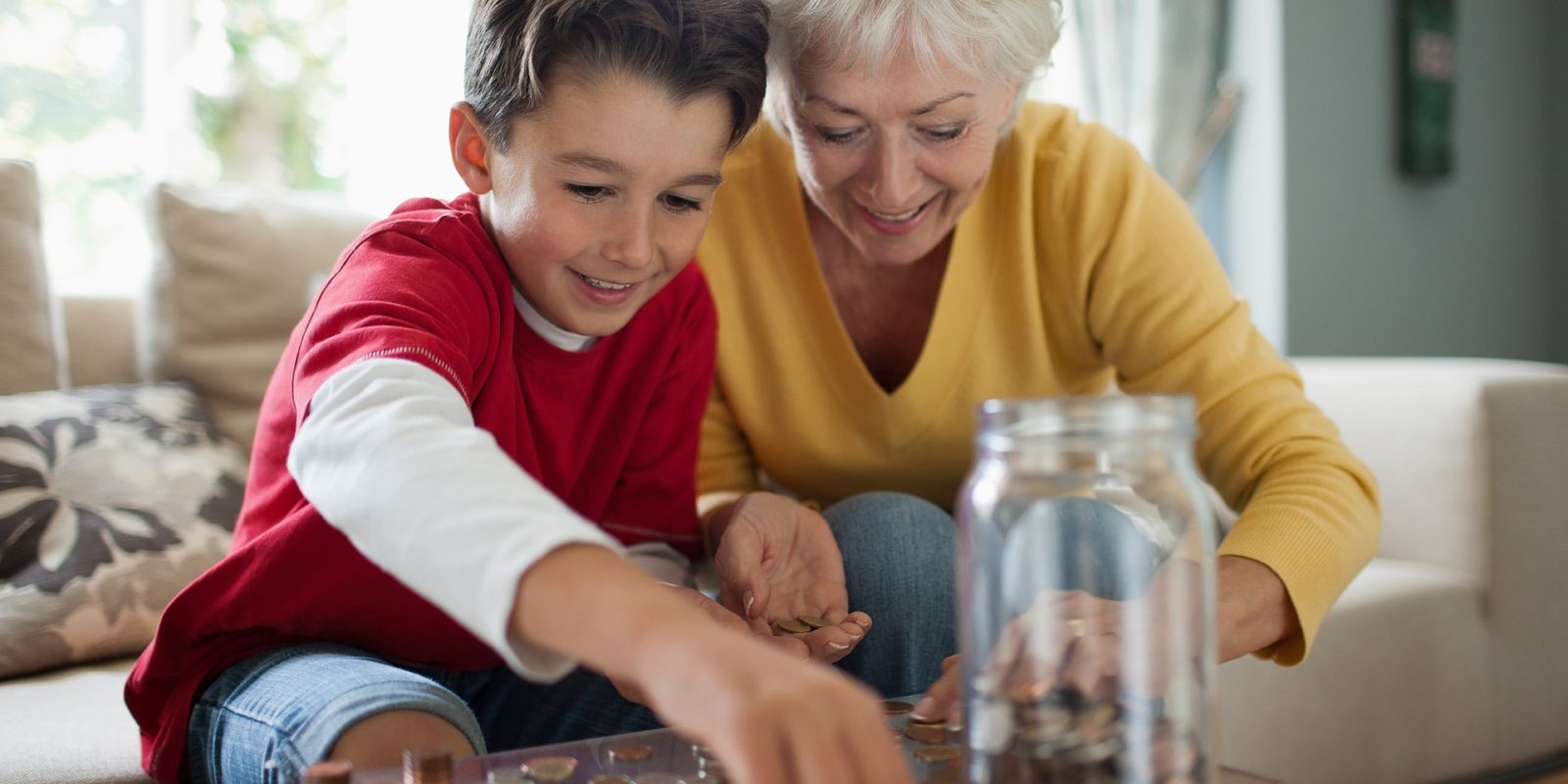 Young boy in red shirt and older woman in yellow shirt count coins on a table with a jar full of coins on it