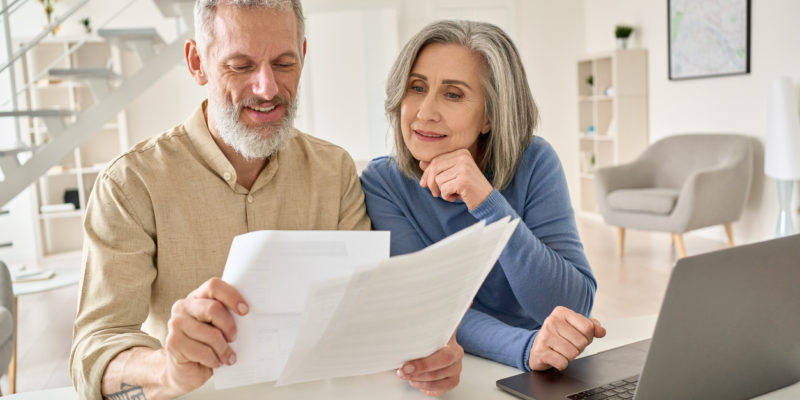 older man and woman sit at counter and look at sheets of paper in front of laptop