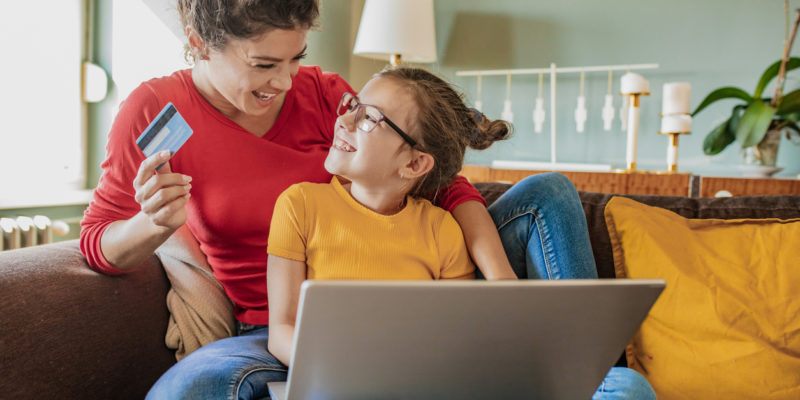 woman in red shirt holds credit card and young girl in a yellow shirt sits on her lap and holds a computer