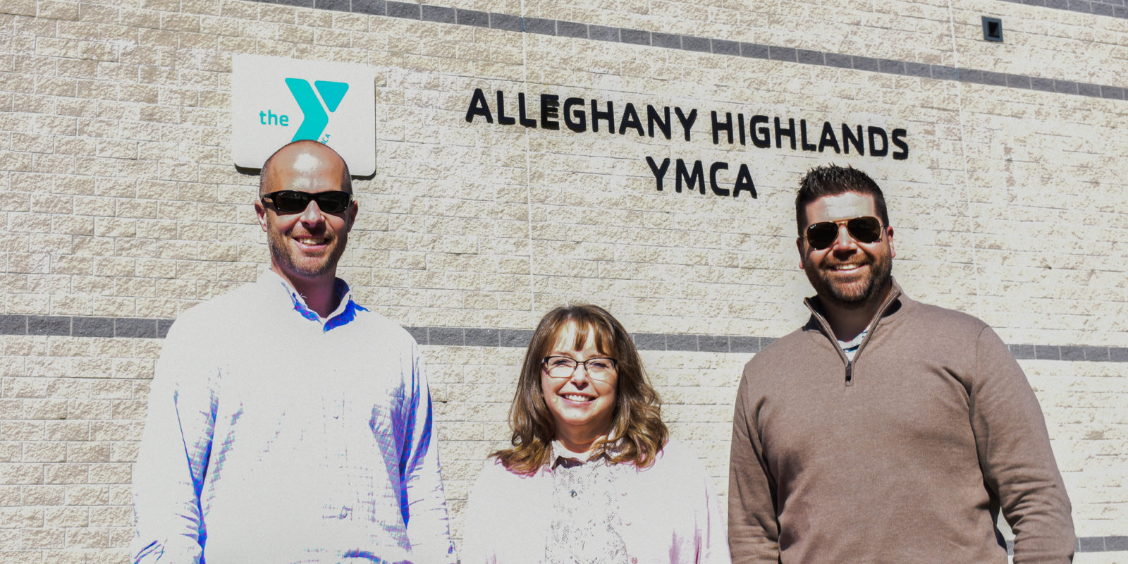 Two men stand on either side of a woman, standing in front of Alleghany Highlands YMCA building